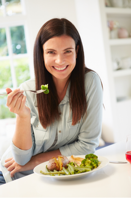Photo of a woman with long brown hair sitting down at a table with a plate of food in front of her. She is holding a fork of food up to her mouth and smiling. 